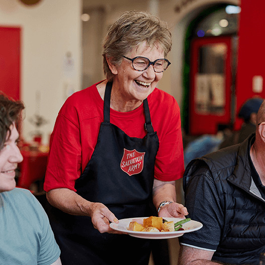 Salvation Army volunteer smiling as she serves a warm meal to guests at a community centre.