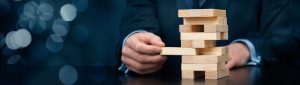 A close-up of a wooden block tower with a single block being carefully removed, illustrating the impact that hidden risks or weak points can have on business stability, continuity and operational resilience.